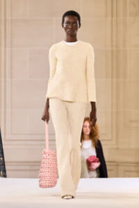 Model walks the runway in a cream textured knit sweater and matching wide-leg pants, carrying a pink woven tote bag in her left hand.