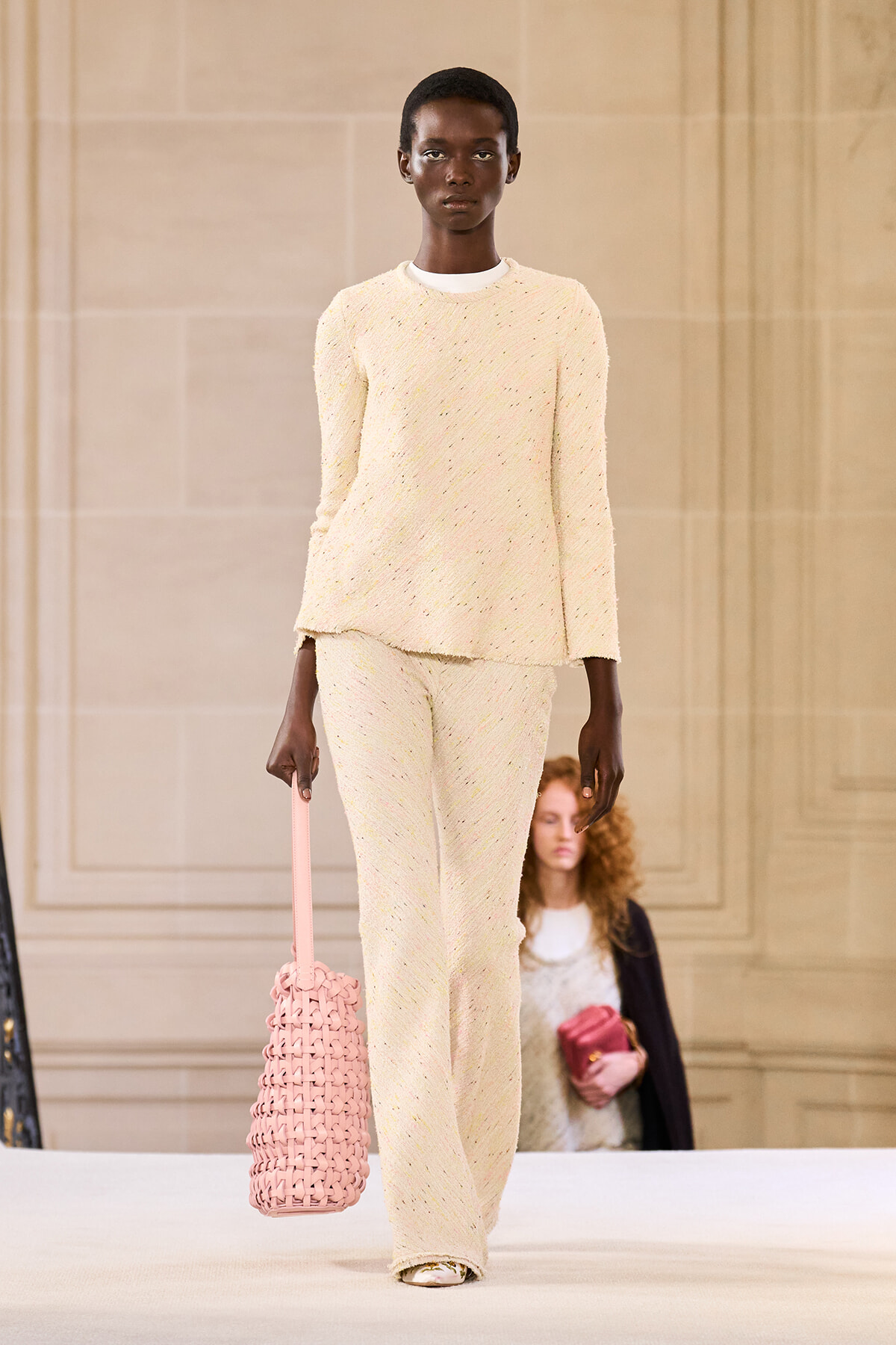 Model walks the runway in a cream textured knit sweater and matching wide-leg pants, carrying a pink woven tote bag in her left hand.
