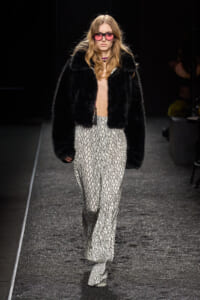 Model walks runway wearing a black fur jacket over a patterned wide-leg jumpsuit, pink sunglasses, and a purple choker.