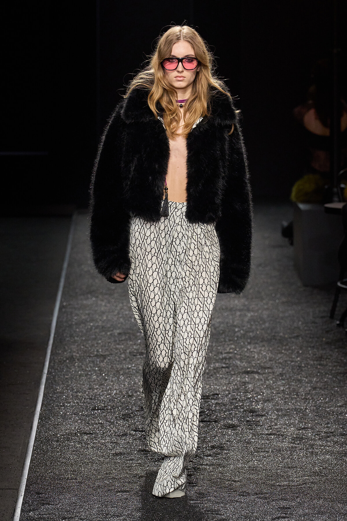 Model walks runway wearing a black fur jacket over a patterned wide-leg jumpsuit, pink sunglasses, and a purple choker.