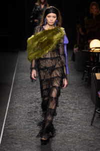Model walks the runway wearing a sheer black beaded gown with a lime-green faux fur stole draped over one shoulder and bold blue accessories.