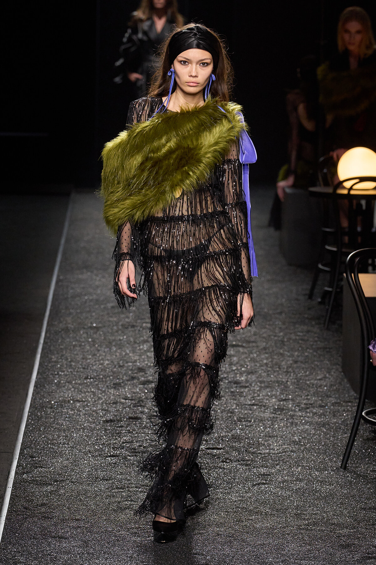 Model walks the runway wearing a sheer black beaded gown with a lime-green faux fur stole draped over one shoulder and bold blue accessories.