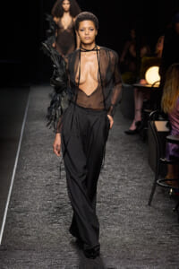 Model walks a fashion runway in a sheer black blouse with a deep neckline, black trousers, and a large feathered arm accessory on the left.