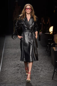 Model walking the runway in a black leather trench with a belt, pink sunglasses, and red-strap heels while carrying a black bag on her left arm