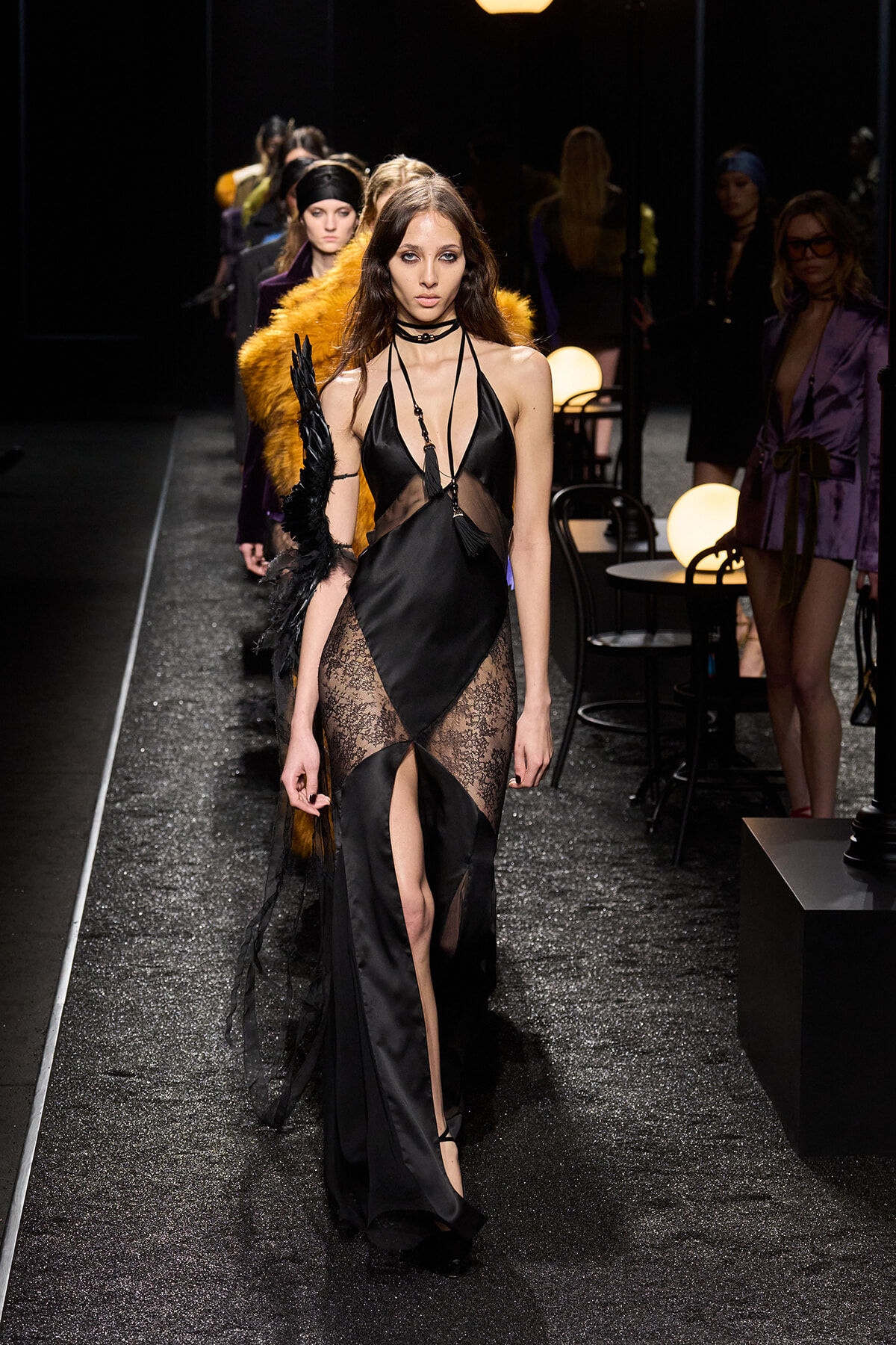 Runway model in a black satin gown with sheer lace panels, high slit, and long gloves, walking the catwalk amidst spectators.