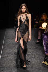 Model walking the runway in a black satin slip dress with sheer lace panels and layered tassel necklace accessories.