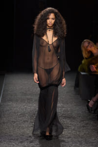 Model on a runway wearing a sheer black dress with visible lingerie and layered necklaces.
