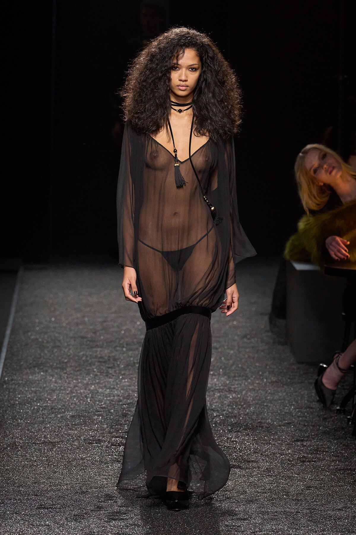Model on a runway wearing a sheer black dress with visible lingerie and layered necklaces.