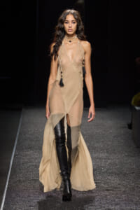 Model walking a runway in a sheer beige halter dress with long tassel necklaces and black thigh-high boots.