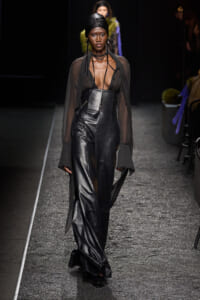 Model walks a fashion runway in a sheer black blouse over a black leather corset and wide-leg pants, with a black head wrap and layered necklaces.