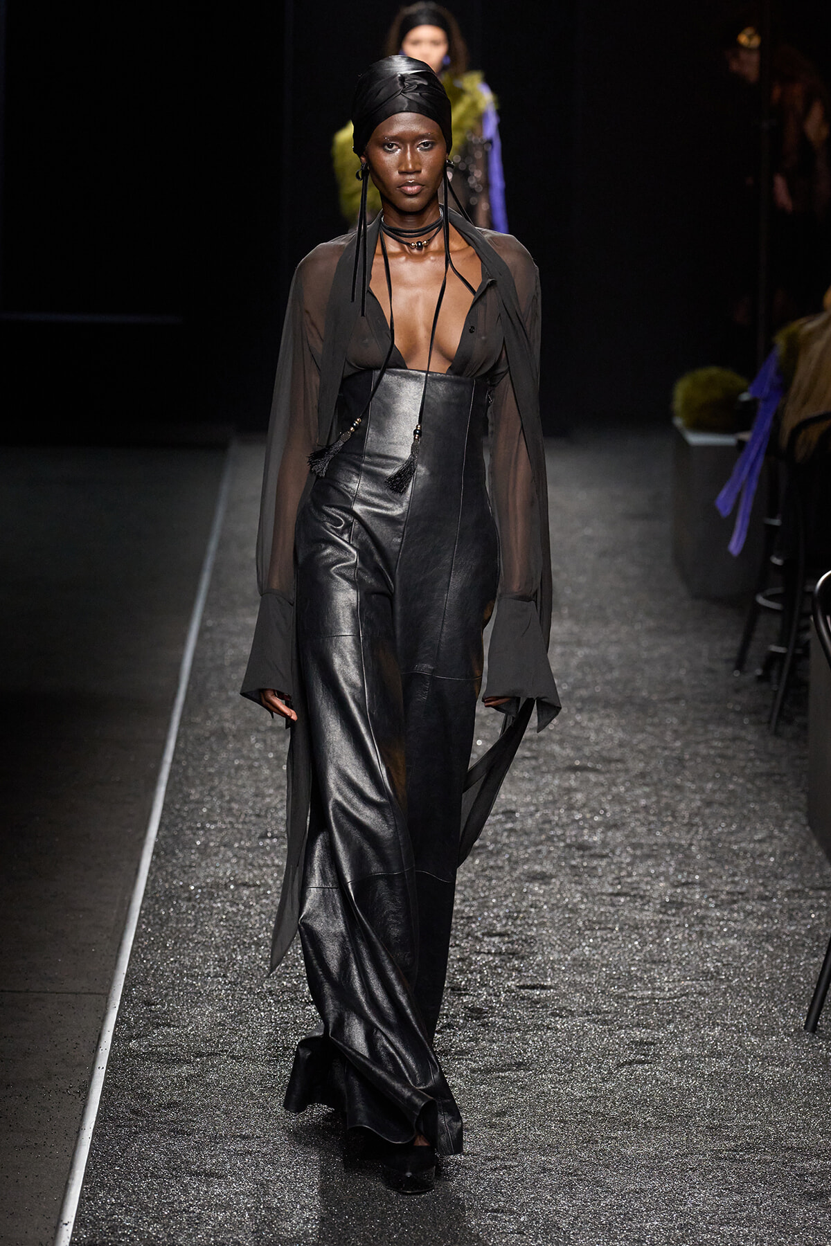 Model walks a fashion runway in a sheer black blouse over a black leather corset and wide-leg pants, with a black head wrap and layered necklaces.