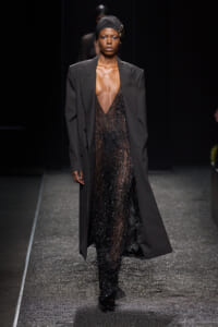 Runway model wearing a tailored black blazer over a sheer, beaded black gown with a black headwrap and gold brooch walking the catwalk.