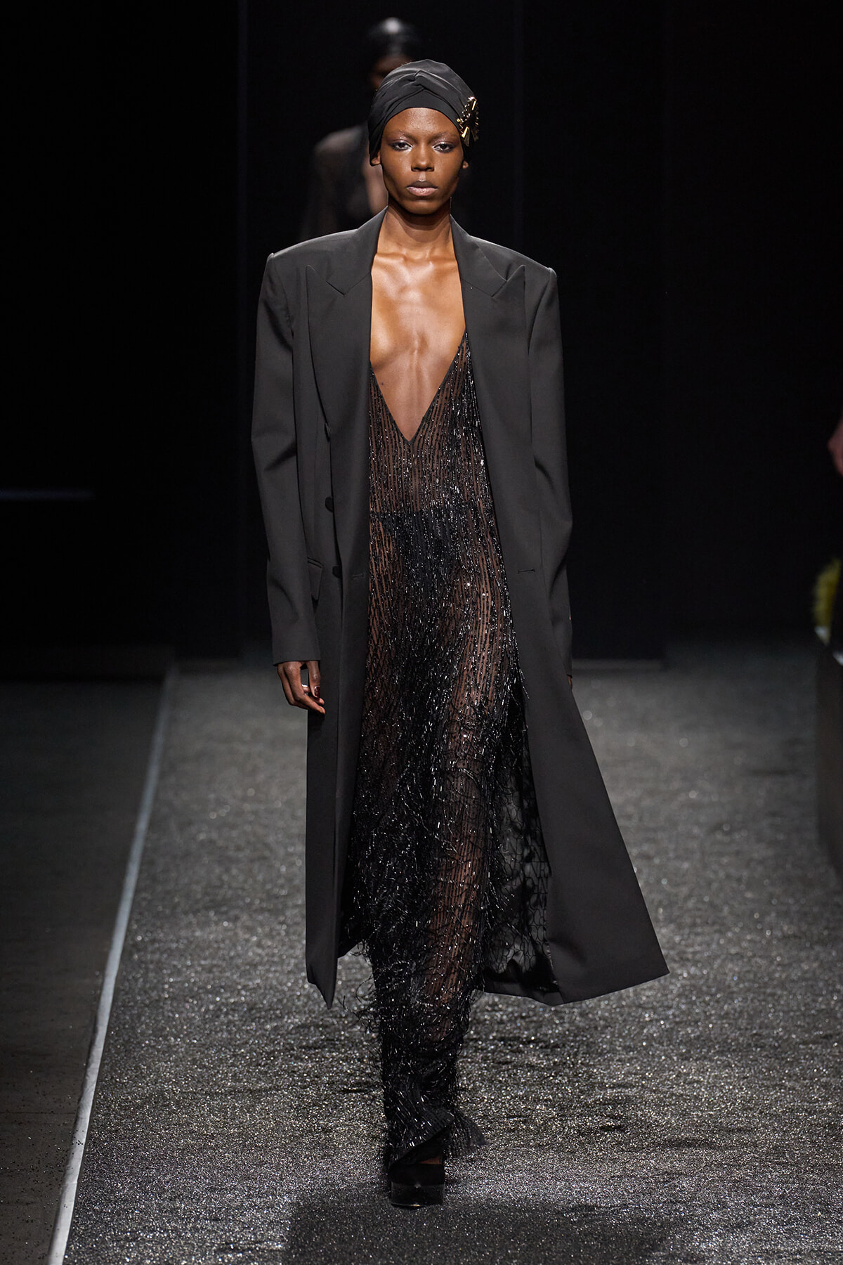 Runway model wearing a tailored black blazer over a sheer, beaded black gown with a black headwrap and gold brooch walking the catwalk.