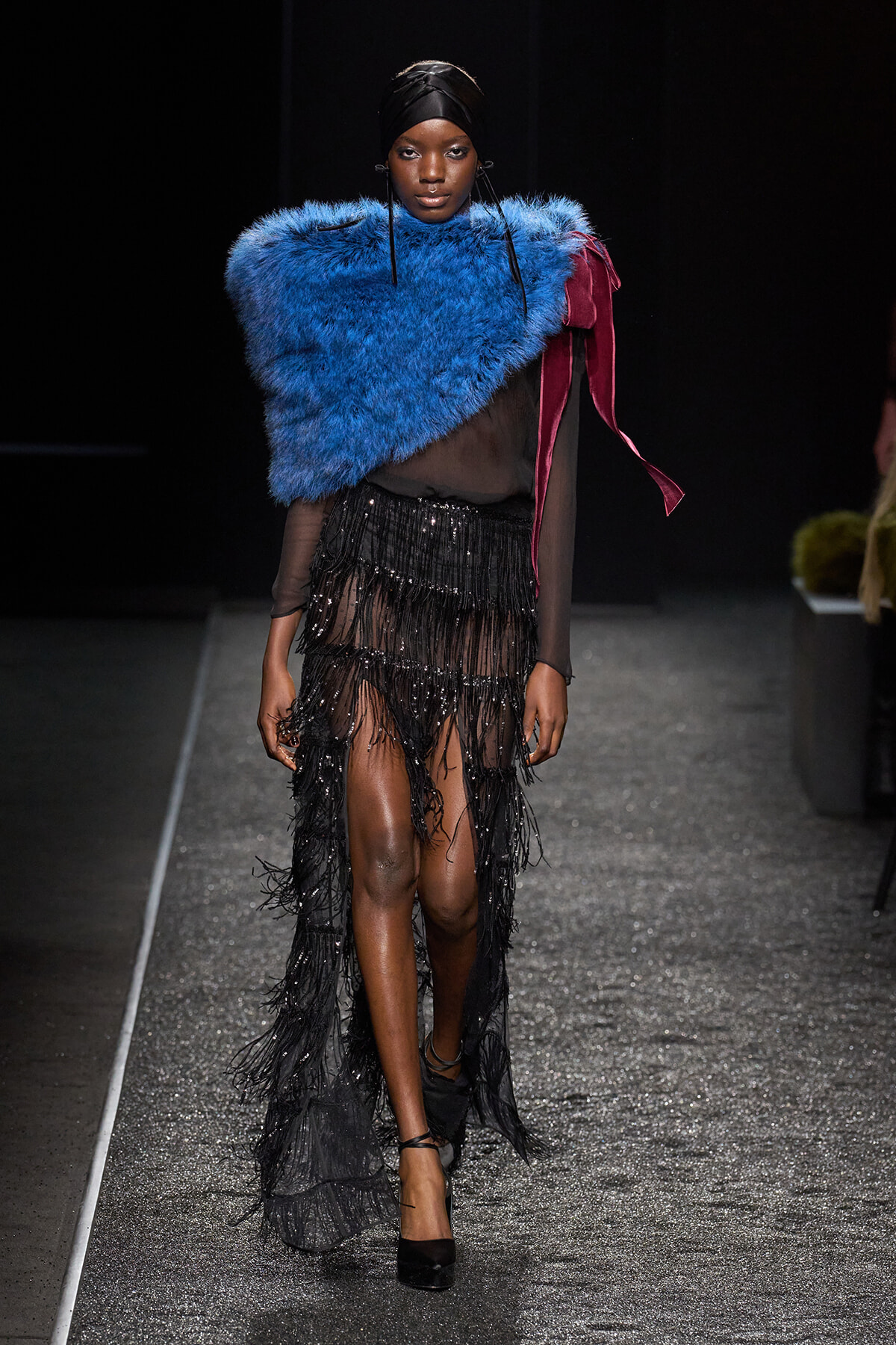 Model walking the runway in a vivid blue faux-fur shawl over a sheer black top and black fringe skirt.