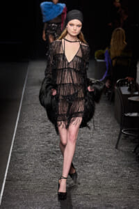 Model walks the runway in a sheer black beaded dress with fringe, black headscarf, and a faux fur wrap held at sides on a glittery catwalk.