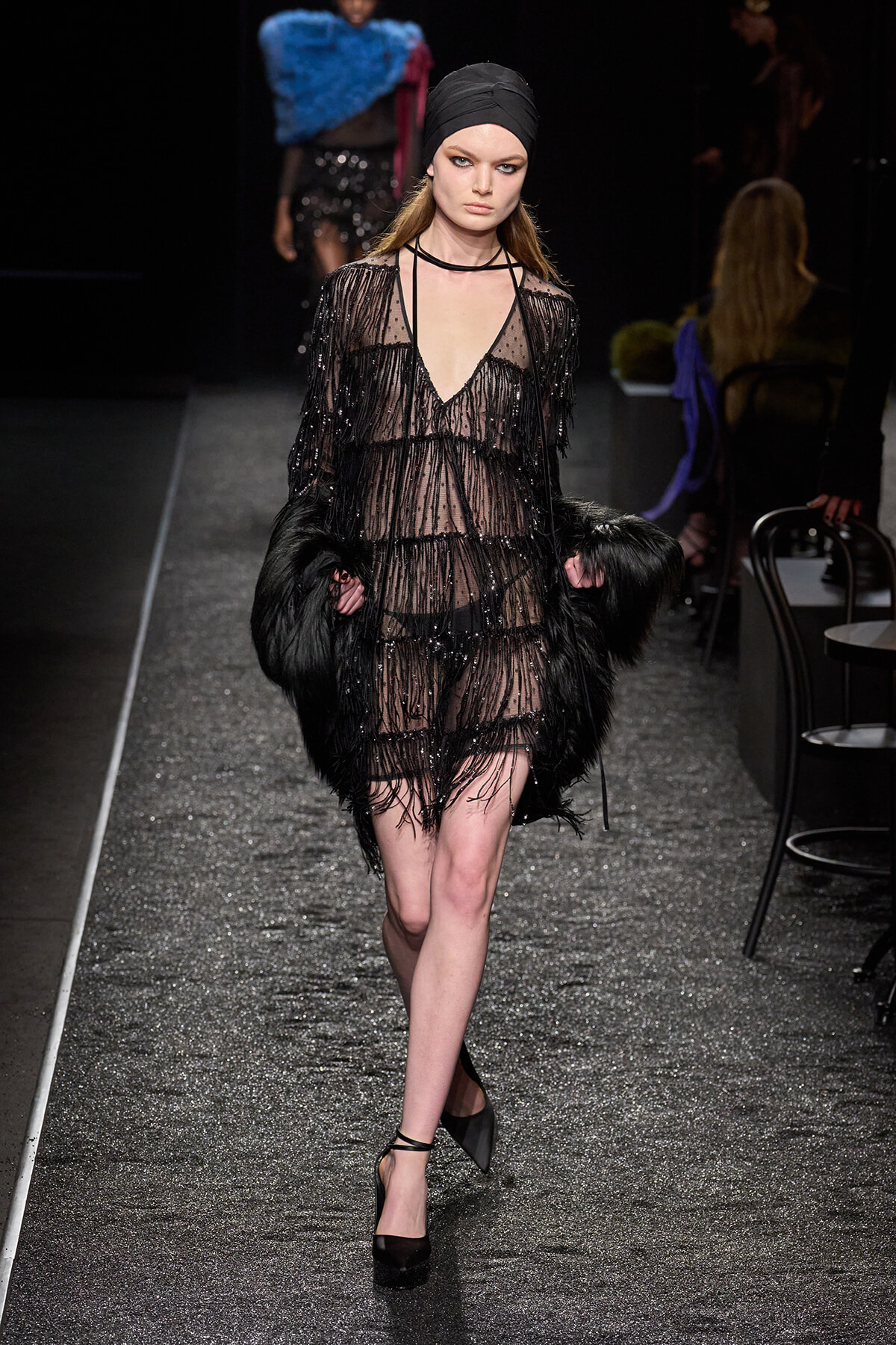 Model walks the runway in a sheer black beaded dress with fringe, black headscarf, and a faux fur wrap held at sides on a glittery catwalk.