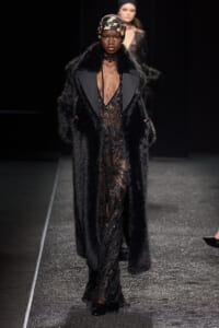 Model walks the runway wearing a long black fur coat over a sheer sequined gown with a headwrap, on a glittery catwalk.