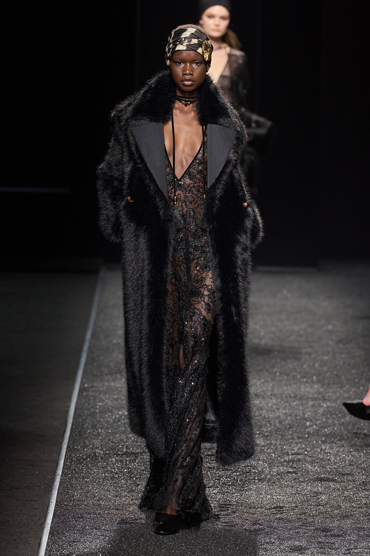 Model walks the runway wearing a long black fur coat over a sheer sequined gown with a headwrap, on a glittery catwalk.