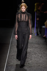 Model walks a runway in a sheer black blouse tucked into pinstriped high-waisted wide-leg pants.