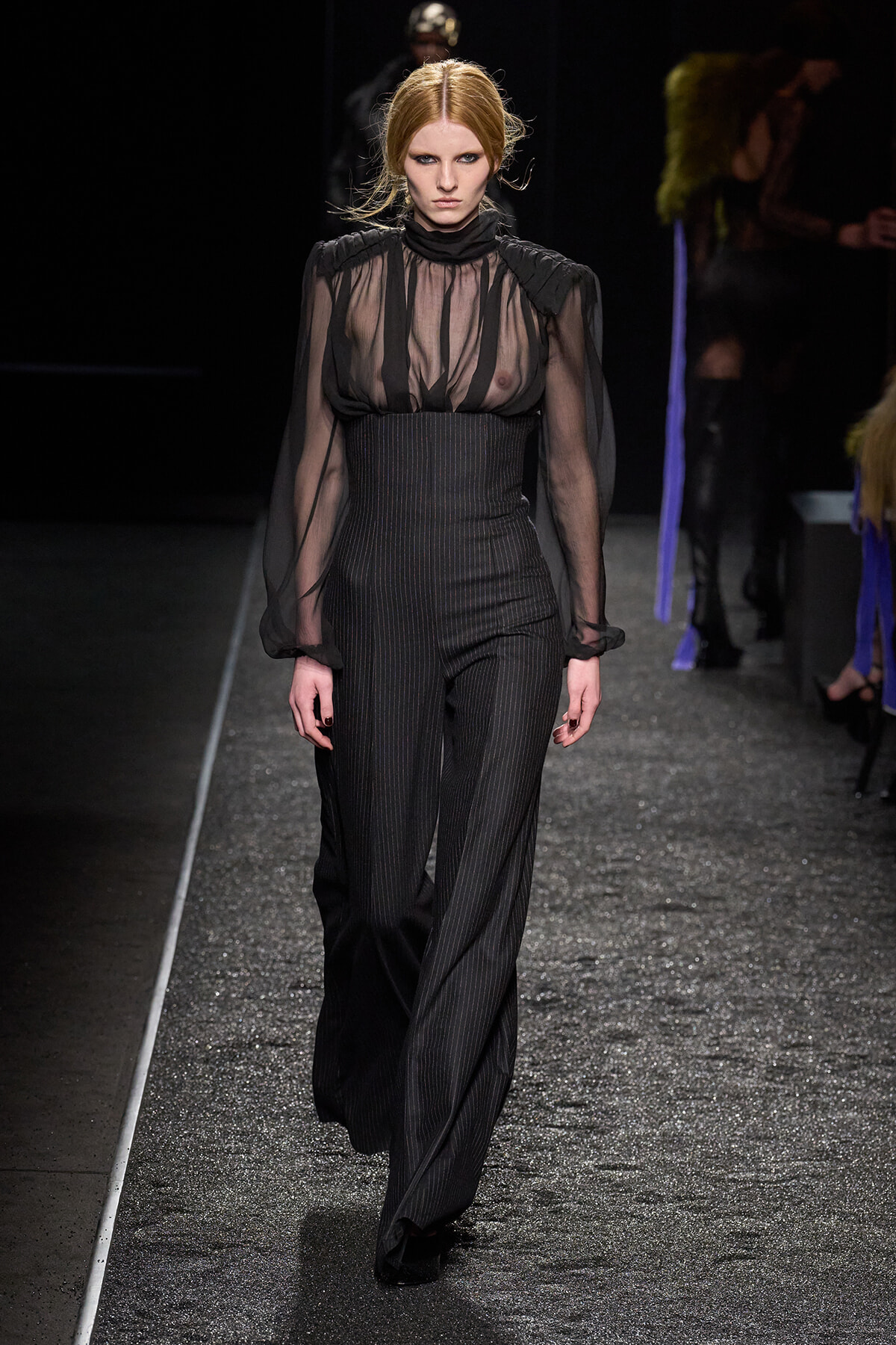 Model walks a runway in a sheer black blouse tucked into pinstriped high-waisted wide-leg pants.