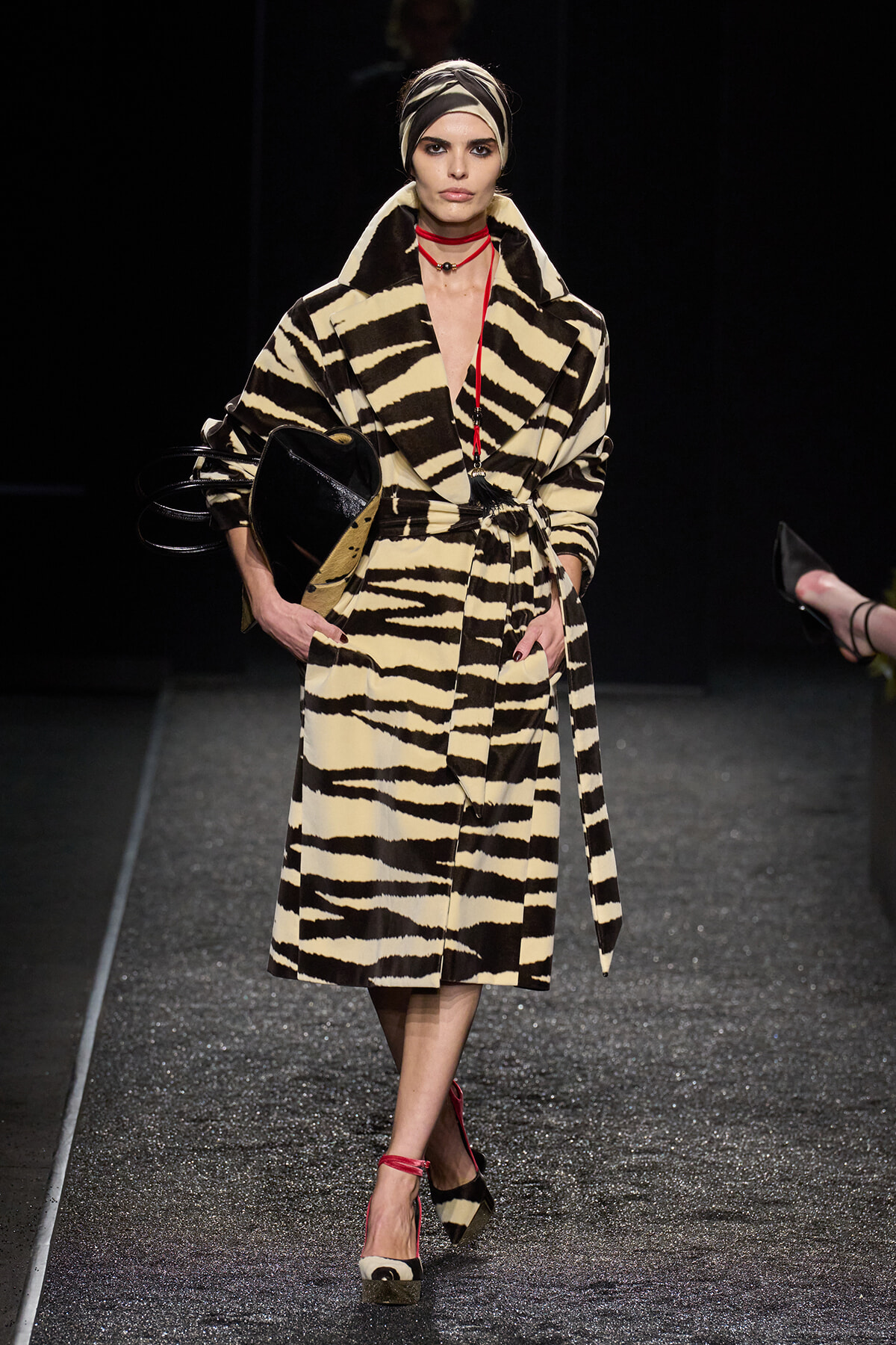 Model walking the runway in a cream-and-black zebra-striped coat, belted at the waist, with a head wrap and red accessories.