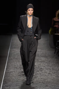 Model walking a dimly lit runway in a black oversized suit with broad-shouldered blazer and wide-leg trousers, black head wrap, and blue beaded tassel necklaces.