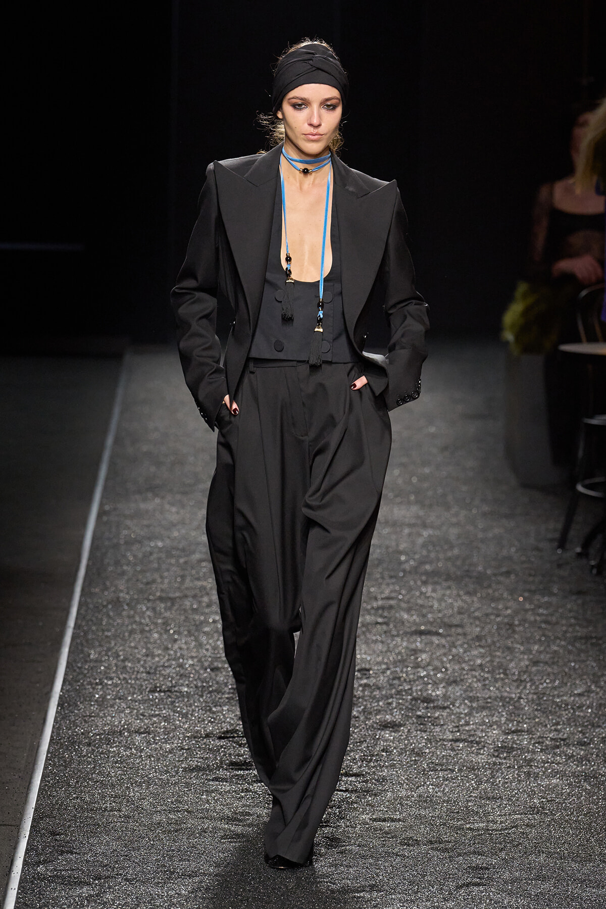 Model walking a dimly lit runway in a black oversized suit with broad-shouldered blazer and wide-leg trousers, black head wrap, and blue beaded tassel necklaces.