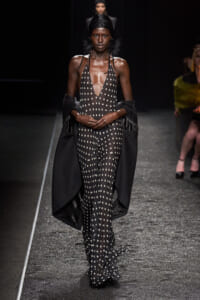Model walks the runway in a black polka-dot halter jumpsuit with a dramatic black cape draped over her arms.