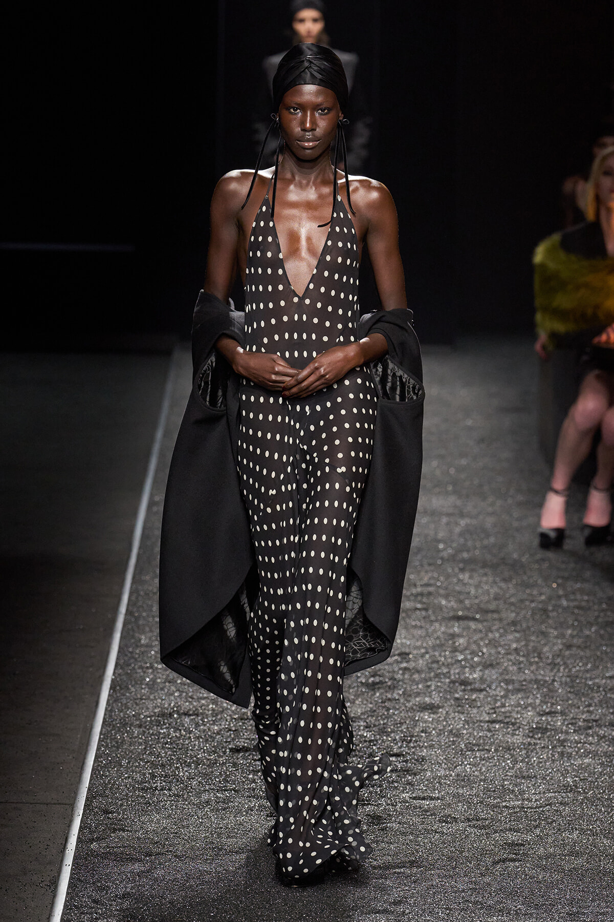 Model walks the runway in a black polka-dot halter jumpsuit with a dramatic black cape draped over her arms.