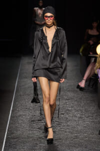 Model walks a black satin mini dress with deep V neckline, head wrap, pink sunglasses, and layered tassel necklaces on a glittery runway, carrying a small black handbag.