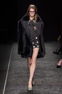 Model walks a glittery runway in a black fur coat over a sheer polka-dot dress with beige platform heels and pink-tinted sunglasses.