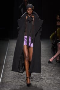 Model on a runway wearing a long black oversized coat, sheer black blouse, purple shorts, and a black headwrap with a gold brooch.