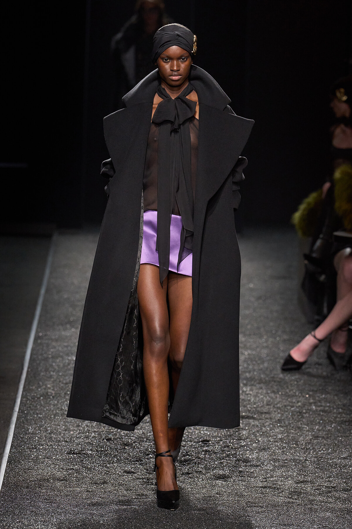 Model on a runway wearing a long black oversized coat, sheer black blouse, purple shorts, and a black headwrap with a gold brooch.