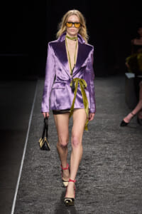 Model walking the runway in a shiny purple blazer dress with a lime sash, yellow-tinted sunglasses, and black-and-beige espadrille heels, carrying a small black and beige handbag.