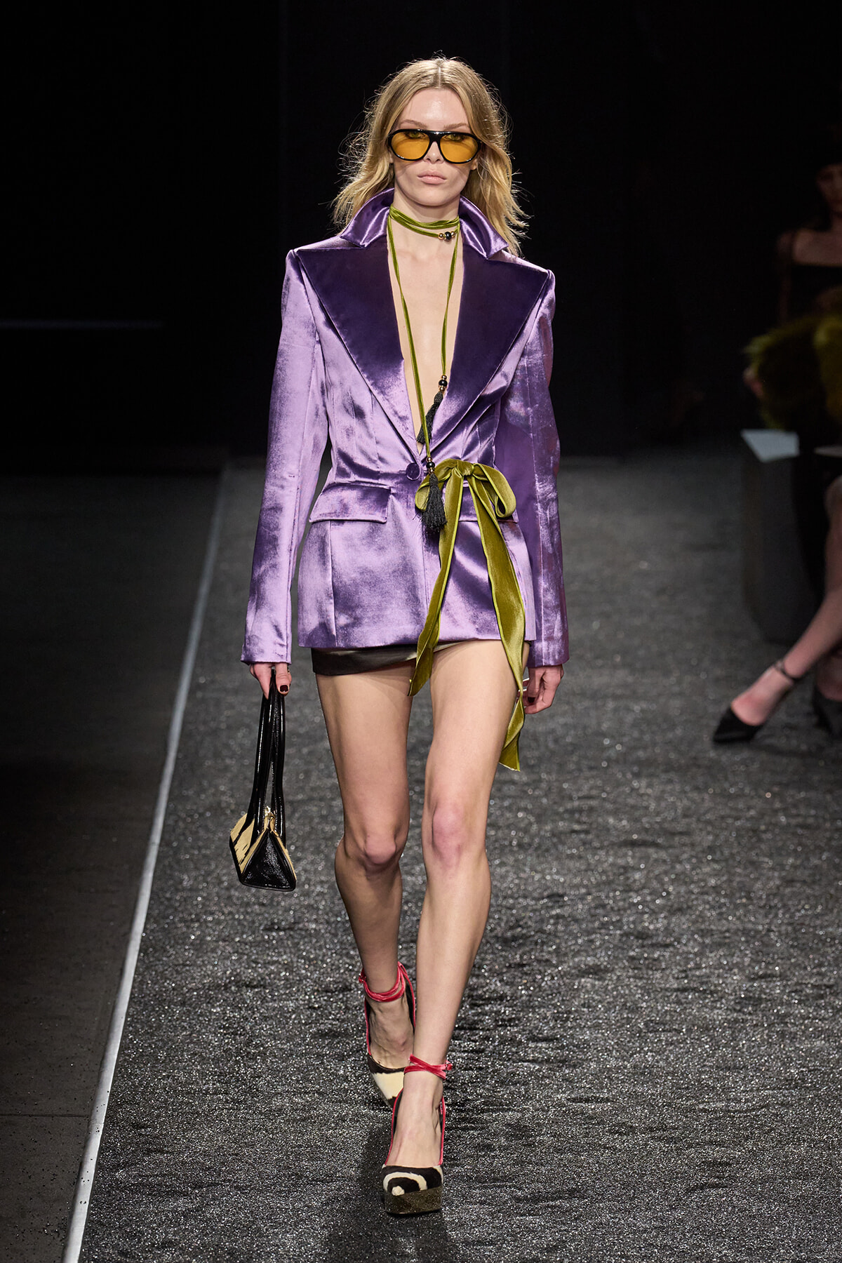 Model walking the runway in a shiny purple blazer dress with a lime sash, yellow-tinted sunglasses, and black-and-beige espadrille heels, carrying a small black and beige handbag.