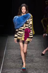Model on a runway wearing a zebra-print blazer, blue fur stole, beige dress with lace hem, magenta bow, and color-block heels.