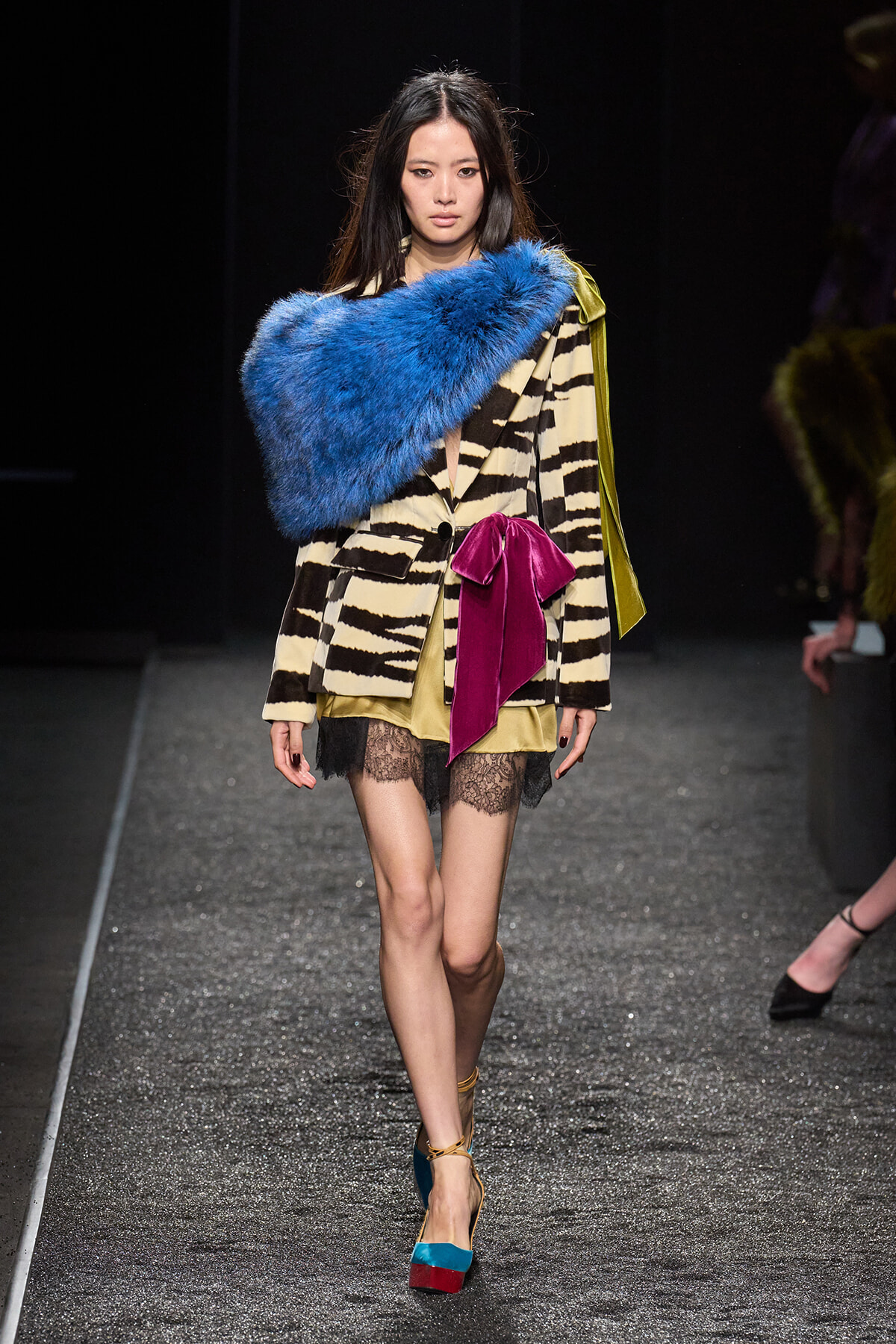 Model on a runway wearing a zebra-print blazer, blue fur stole, beige dress with lace hem, magenta bow, and color-block heels.