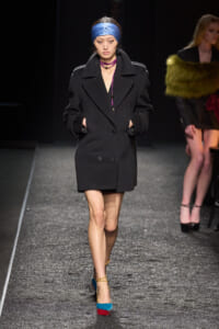 Model walking the runway in a black double-breasted coat, blue headscarf, and color-block teal and red platform heels.