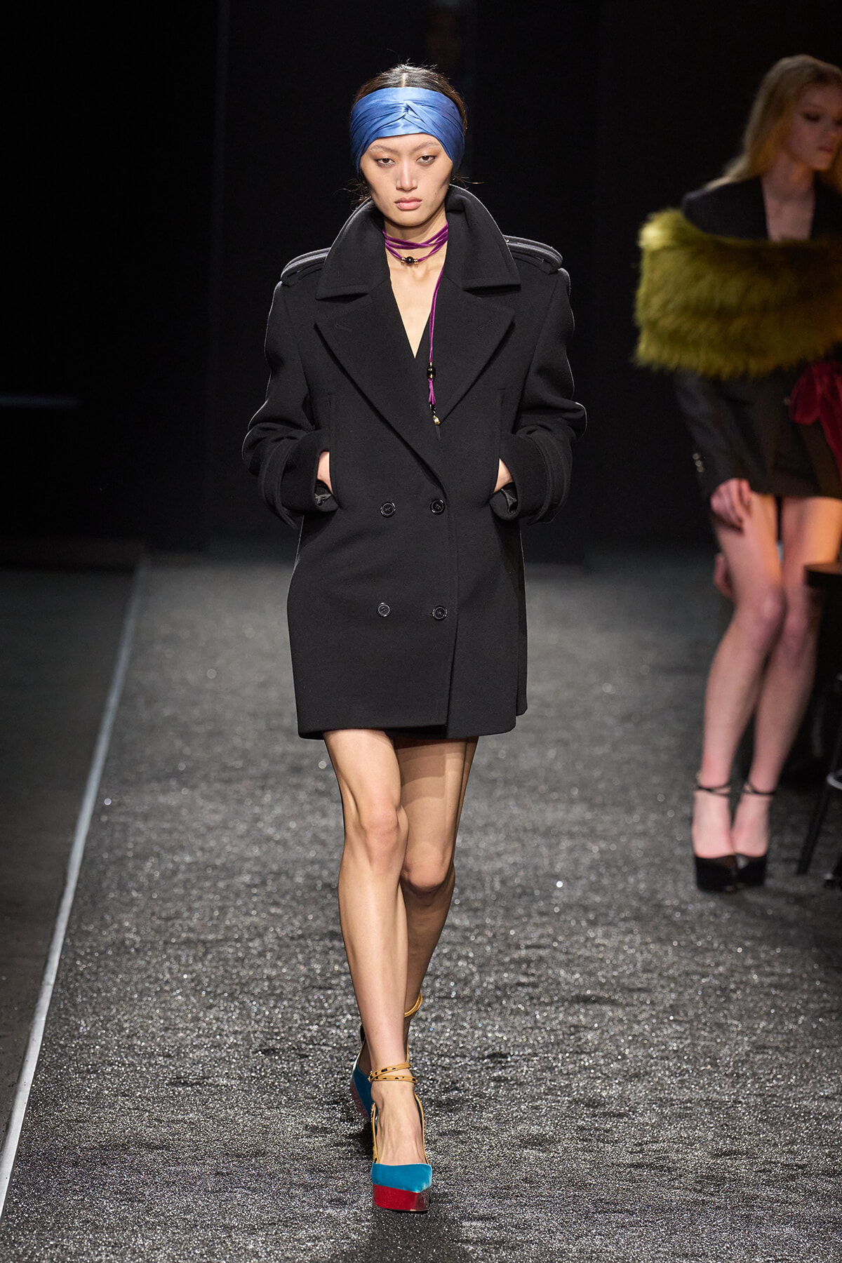 Model walking the runway in a black double-breasted coat, blue headscarf, and color-block teal and red platform heels.