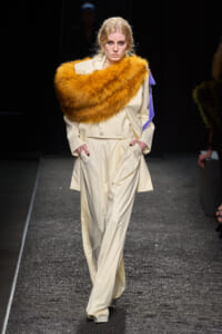 Model walking the runway in a pale cream suit with a large orange fur stole draped over her shoulders and a lavender ribbon on the sleeve, hands in pockets.