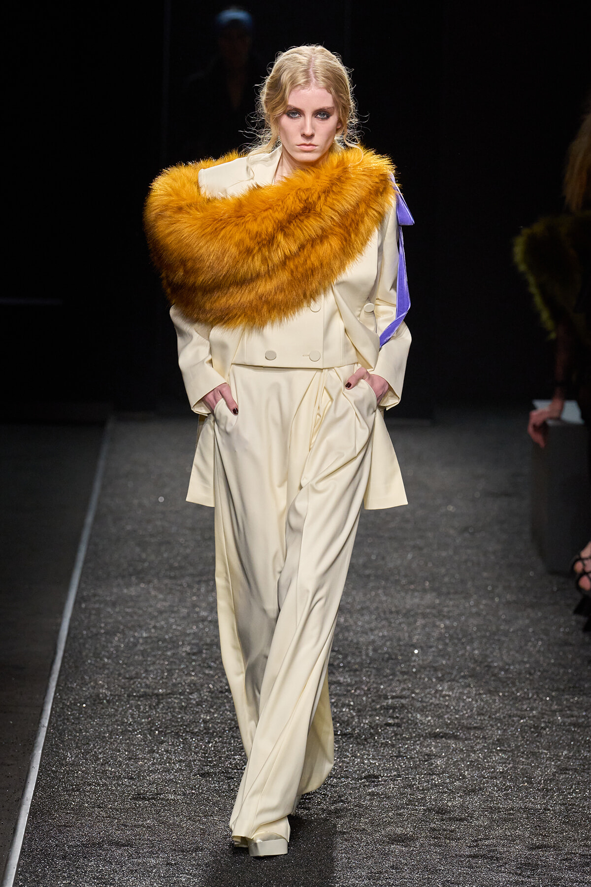 Model walking the runway in a pale cream suit with a large orange fur stole draped over her shoulders and a lavender ribbon on the sleeve, hands in pockets.