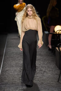Model walks a fashion runway in a sheer beige blouse and high‑waisted black wide-leg pants.