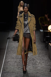 Model walks the runway in a long tan suede trench over a black leather dress, black head wrap, and black heels, carrying a black round handbag.