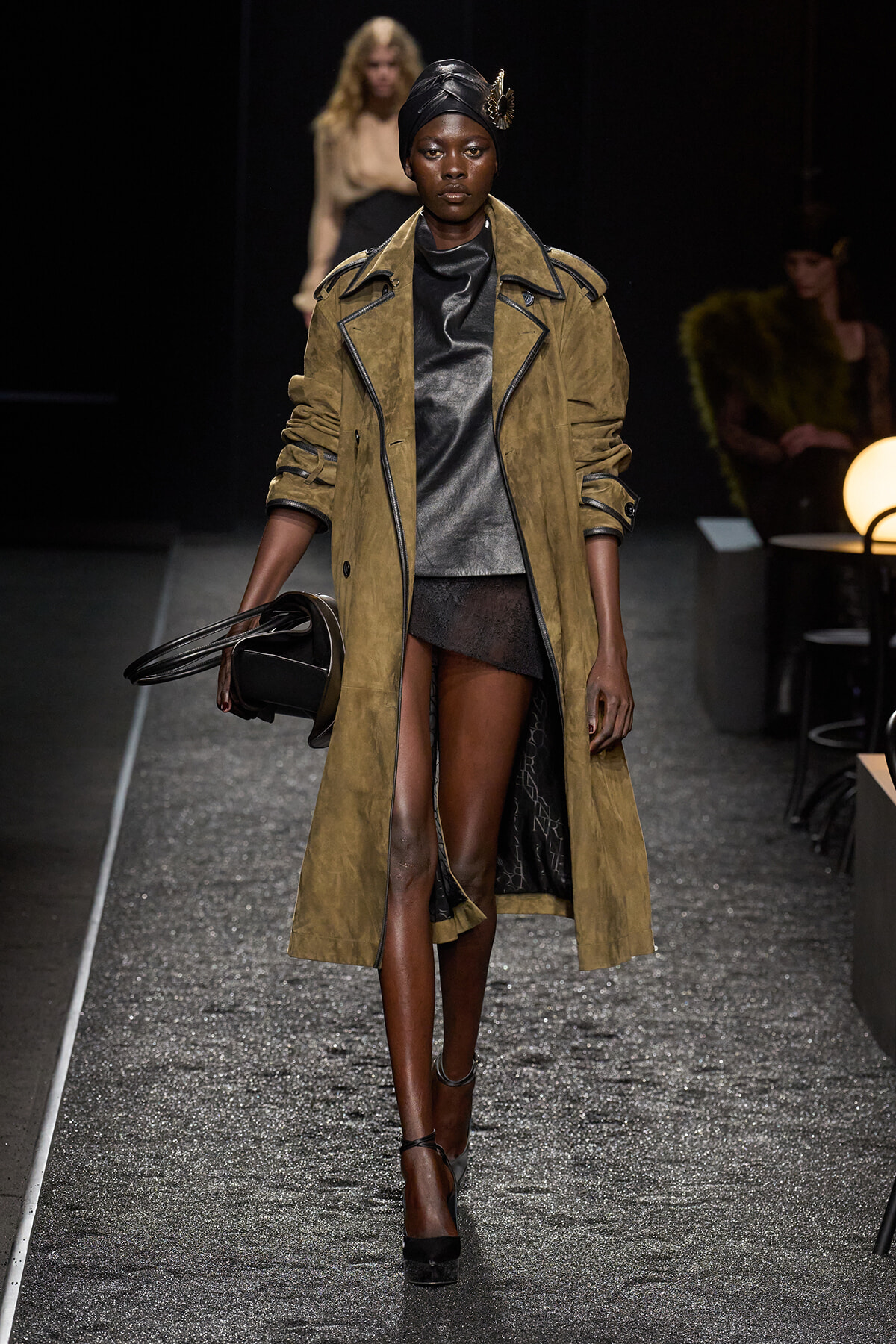 Model walks the runway in a long tan suede trench over a black leather dress, black head wrap, and black heels, carrying a black round handbag.
