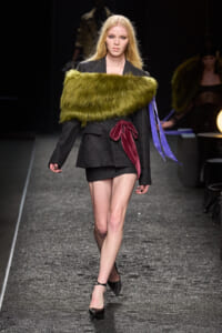 Model walks a runway in a pinstripe blazer-style mini dress with a bright green fur stole and burgundy sash, black heels visible.