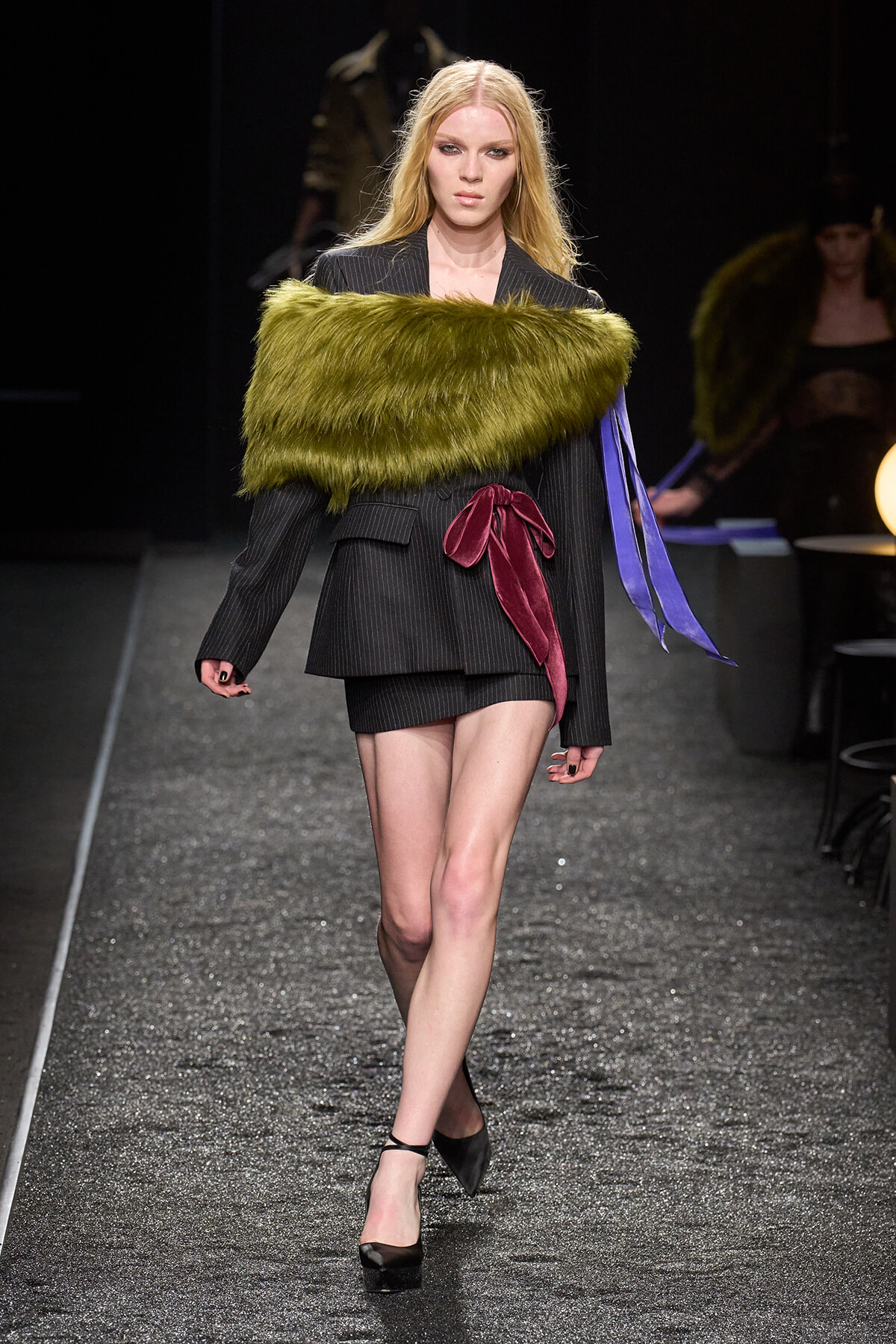 Model walks a runway in a pinstripe blazer-style mini dress with a bright green fur stole and burgundy sash, black heels visible.