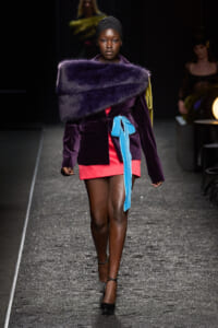 Model wearing a deep purple velvet blazer with a large purple fur stole draped across shoulders, pink mini skirt, and bright blue belt walking a runway in black heels.