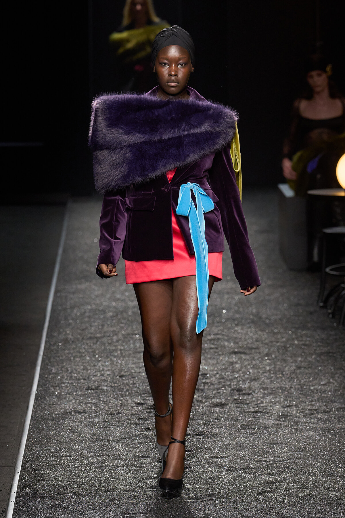 Model wearing a deep purple velvet blazer with a large purple fur stole draped across shoulders, pink mini skirt, and bright blue belt walking a runway in black heels.