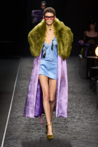 Model on a runway wearing a light blue satin mini dress, a lime-green fur stole, and a long purple velvet coat with chunky sunglasses and gold heels.