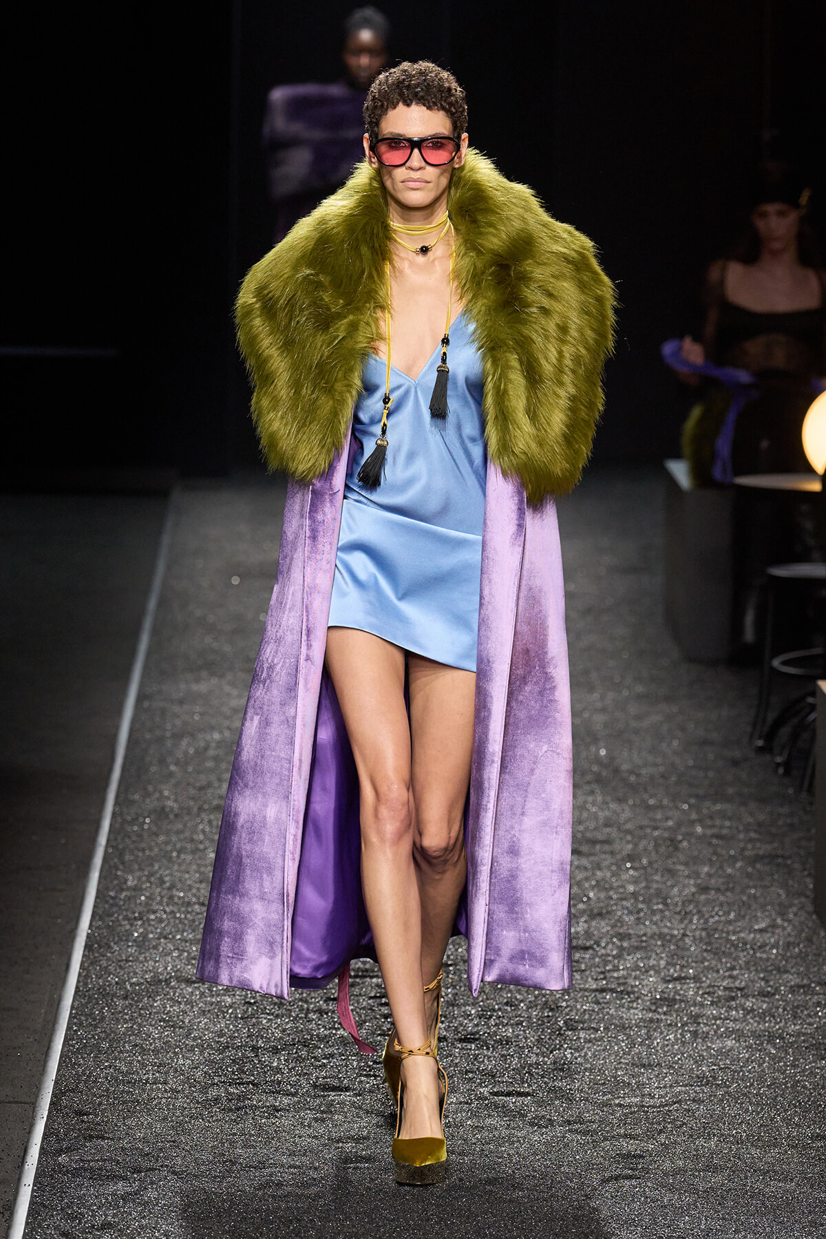 Model on a runway wearing a light blue satin mini dress, a lime-green fur stole, and a long purple velvet coat with chunky sunglasses and gold heels.